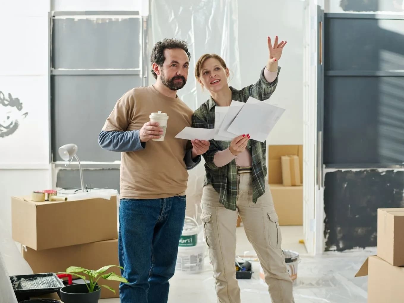 A man holding a coffee cup and a woman holding paint color swatches stand together in a room mid-renovation, with the woman pointing upward as if discussing paint or design choices. Cardboard moving boxes, paint buckets, a potted plant, and plastic-covered furniture and doors fill the space, with protective sheeting also covering the windows in the background.