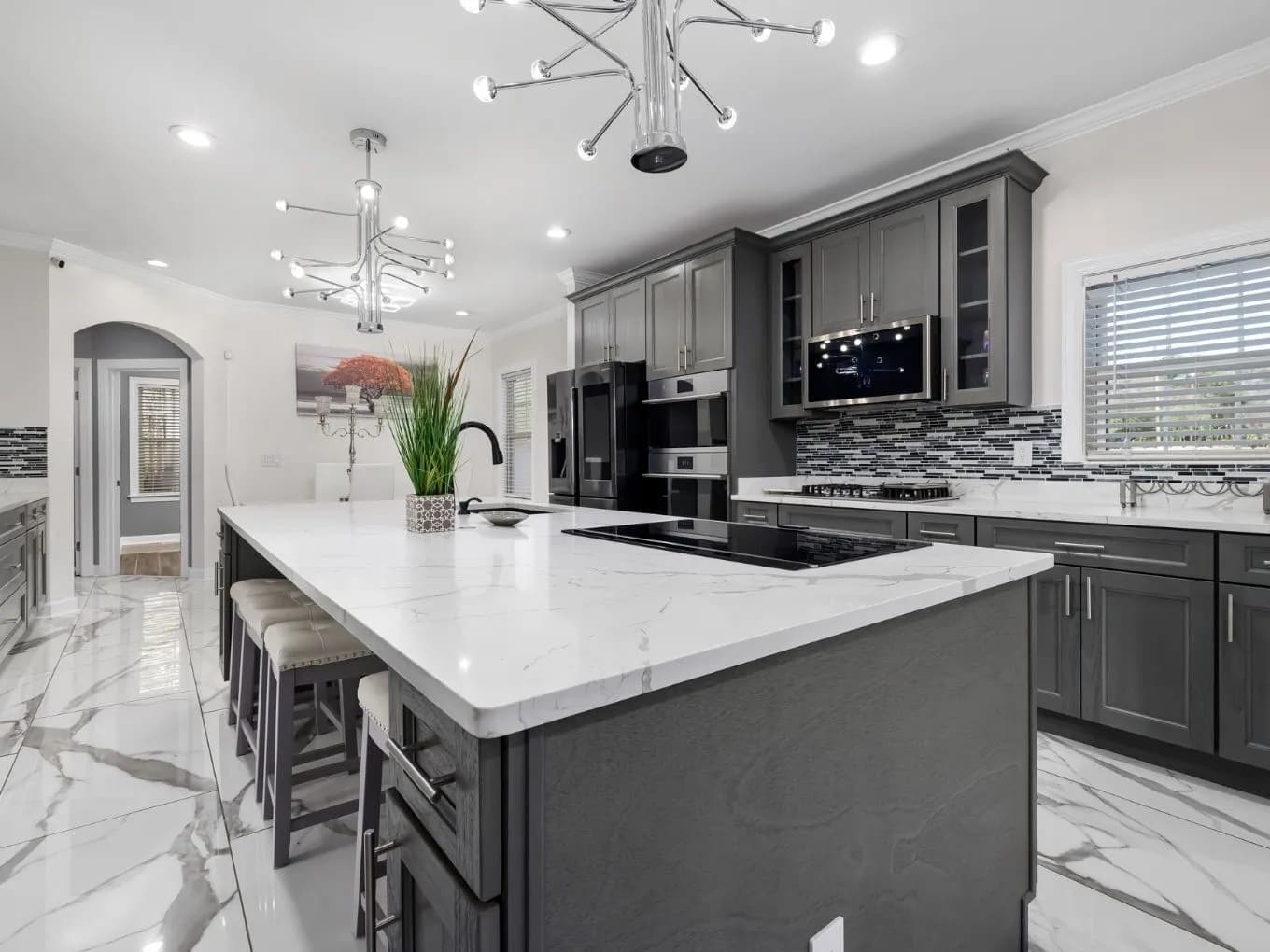 Modern kitchen with a large gray island topped in white marble-look quartz, featuring a built-in cooktop, sink, and upholstered barstools. Gray cabinetry lines the surrounding walls, paired with a mosaic tile backsplash, stainless steel double ovens, microwave, and refrigerator, and glossy marble-patterned floor tile. Two modern chrome chandeliers hang above the island, with an arched doorway and decorative artwork visible in the background hallway.