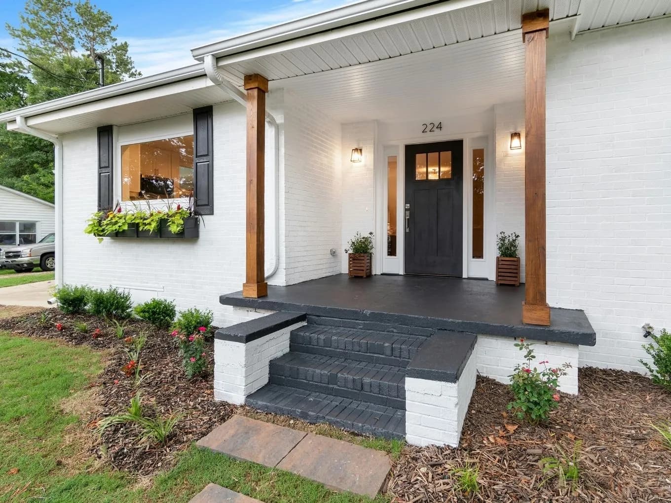 Front entrance of a white brick ranch-style home with the address "224" above a black paneled front door with sidelight windows. The porch has stained wood columns, black wall sconces on either side of the door, and small wood planters with greenery. Black-painted brick steps lead up to the black-finished porch floor, flanked by black shutters on a nearby window with a flower box. Landscaping with mulch, shrubs, and flowering plants lines the front of the house, with a driveway and parked vehicles visible in the background.