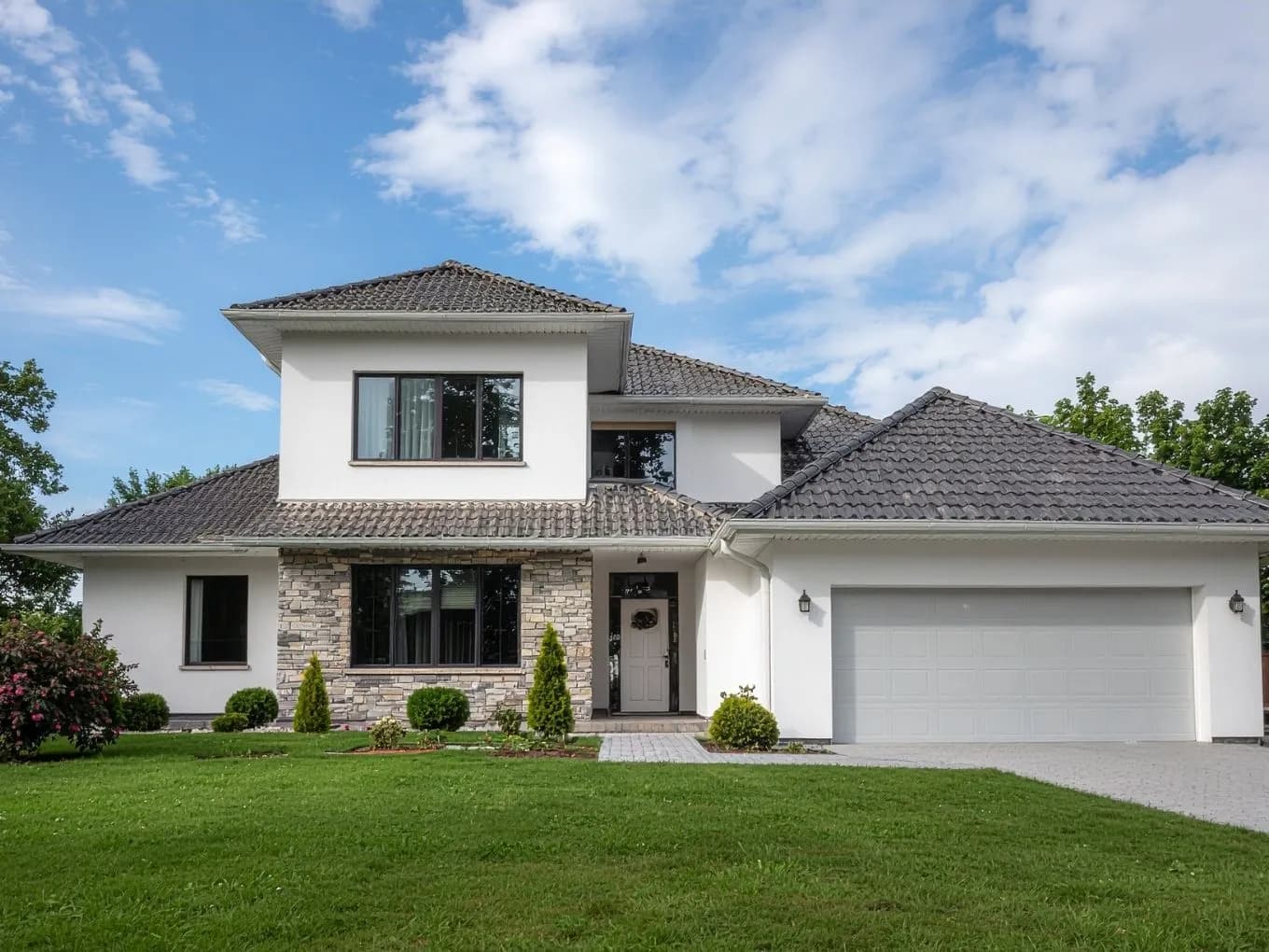 Exterior of a modern two-story white stucco house with dark gray tiled hip roofs and a stone-accented facade near the entrance. A white two-car garage sits to the right of a gray front door, with black-framed windows and small wall lanterns on either side of the entryway. A landscaped front yard with trimmed shrubs, small trees, and a paver walkway and driveway complete the scene under a mostly clear blue sky.