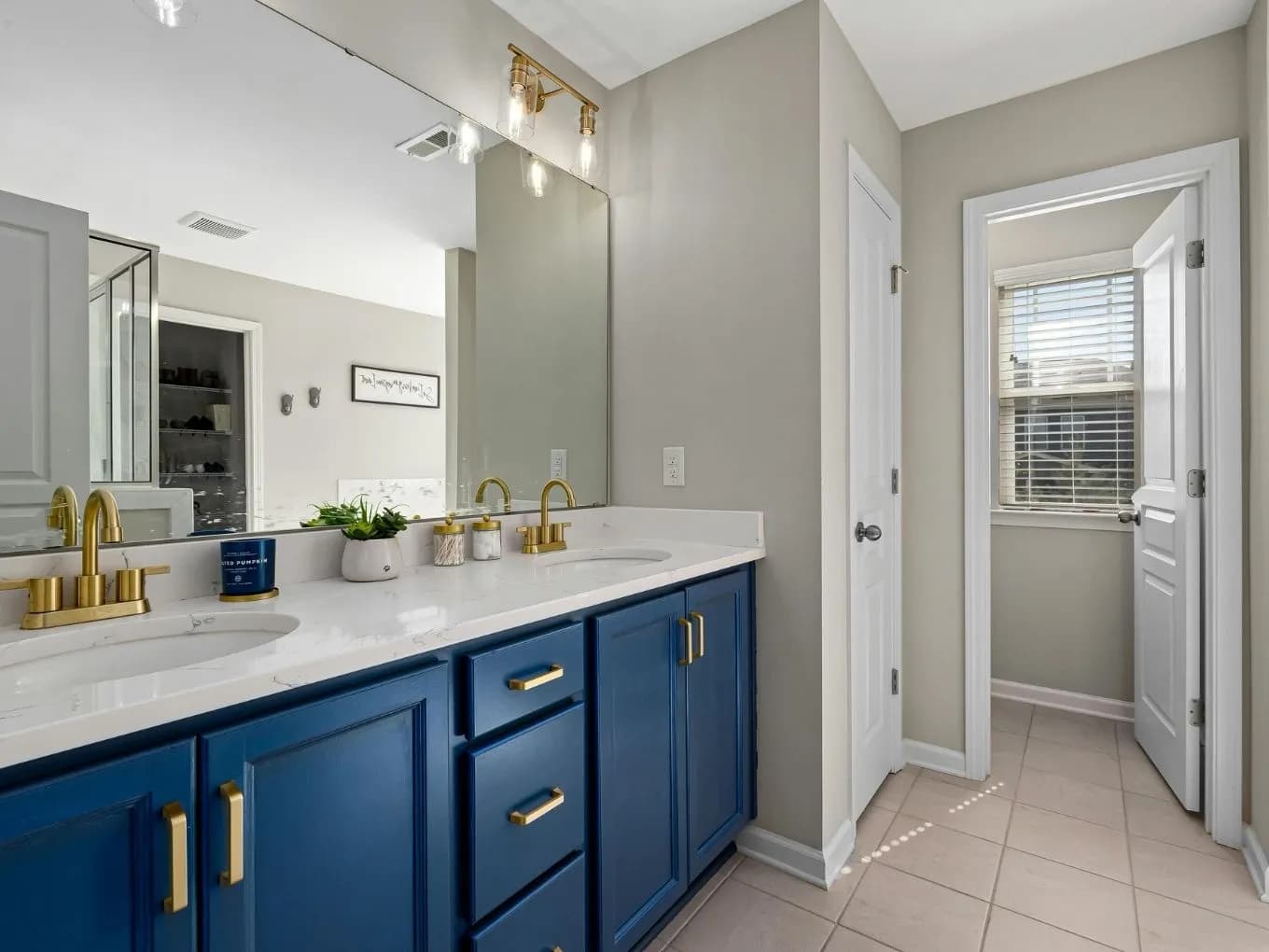 Bathroom with a navy blue double vanity topped in white marble-look quartz, featuring gold faucets, hardware, and a brass vanity light above a large mirror. A small potted plant and decorative jars sit on the counter, with tile flooring extending down a hallway toward a closet door, a window, and an entrance door in the background.