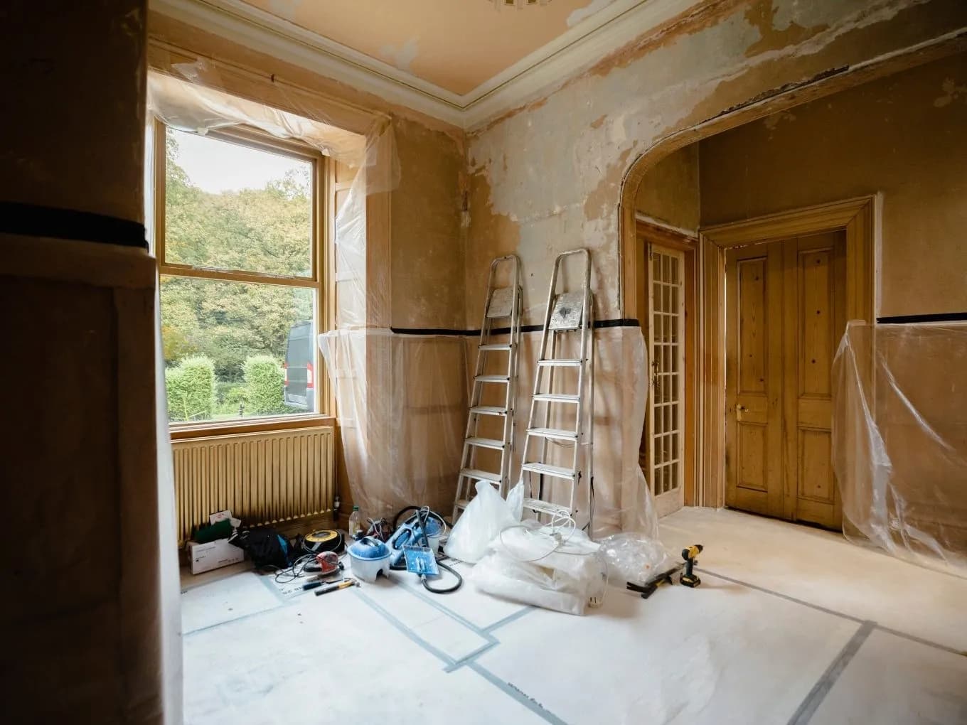 Interior of a room mid-renovation, with walls stripped down to bare plaster and ornate crown molding still intact along the ceiling. Two aluminum step ladders lean against the wall near a large sash window draped in protective plastic sheeting, with a radiator and scattered tools, buckets, and a power drill on the floor below. An arched doorway leads to a hallway with wood-paneled doors, and the floor is covered in protective paper sheeting with visible tape marks.
