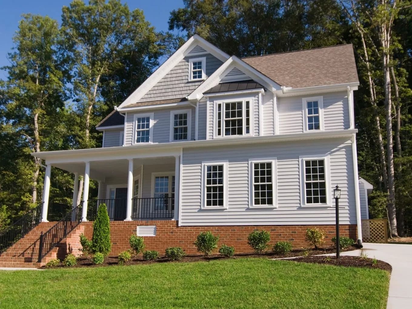 Exterior of a two-story light gray farmhouse-style home with white trim, multiple gables, and a wraparound porch supported by white columns and black metal railings. A red brick foundation lines the base of the house, with newly planted shrubs and mulched beds along the front, and a wide concrete driveway curving toward the right. Tall trees surround the property under a clear blue sky, and a black lamppost stands near the walkway.