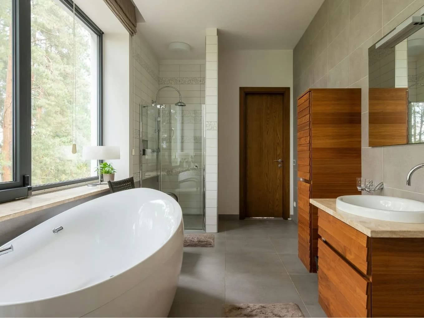 Modern bathroom featuring a freestanding oval white soaking tub positioned beneath a large floor-to-ceiling window overlooking trees, with a glass-enclosed walk-in shower nearby. A dark wood door leads out of the room, and a wood-toned vanity with a round vessel sink and stone countertop sits against a beige tiled wall on the right, with a wood cabinet unit and small bath mats completing the space.
