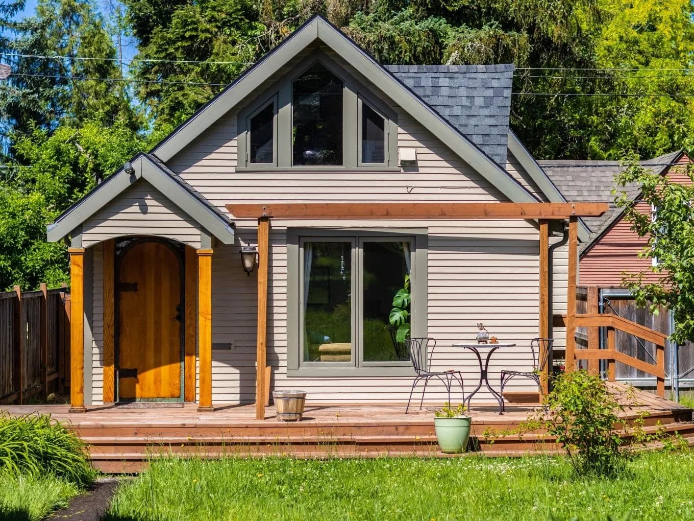 Small cottage-style house with light gray shingle siding, a steep gabled roof, and an arched wooden front door framed by wood posts. A covered wood deck with a pergola extends across the front, furnished with a bistro table, two chairs, and potted plants. Tall trees and greenery surround the yard, with a wooden fence and a neighboring reddish-toned house visible in the background.