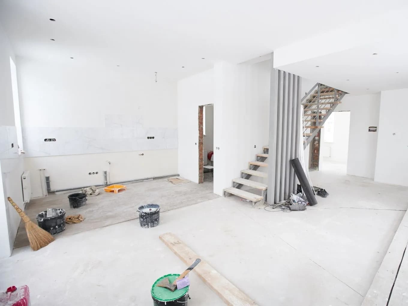 Interior of an open-concept house under construction, with white plastered walls, an unfinished concrete floor, and exposed electrical outlets and plumbing rough-ins along a partially tiled kitchen wall. A concrete staircase with wood tread supports and metal railing leads to an upper level, flanked by vertical decorative slats. Construction supplies including paint buckets, a broom, a trowel, and a wood plank are scattered across the floor, with a small bathroom visible through a doorway in the background.