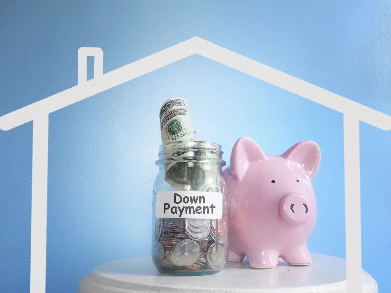 Blueprint of a house as the background. In the front are a pink piggy bank and a jar with dollar bills and coins labeled down payment.