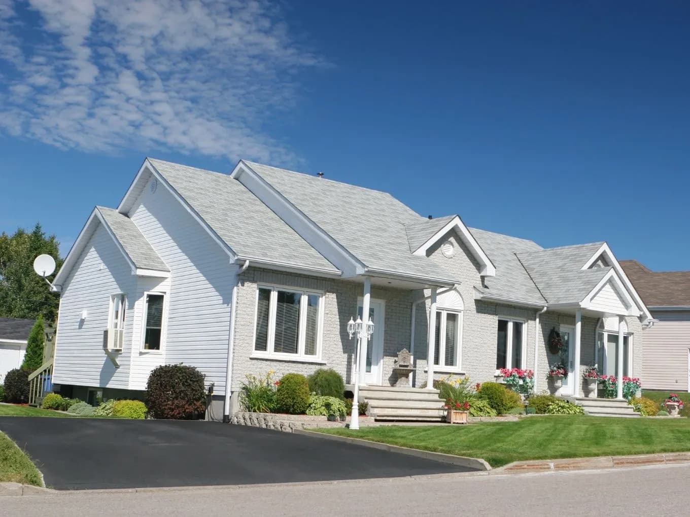 Exterior of a long, single-story ranch-style house with light gray brick and white vinyl siding, featuring multiple gabled rooflines and white-columned entry porches. A decorative lamppost, small water fountain, and colorful flower beds and potted plants decorate the front lawn, with an asphalt driveway curving in from the street. The house extends across a wide lot under a mostly clear blue sky with wispy clouds.