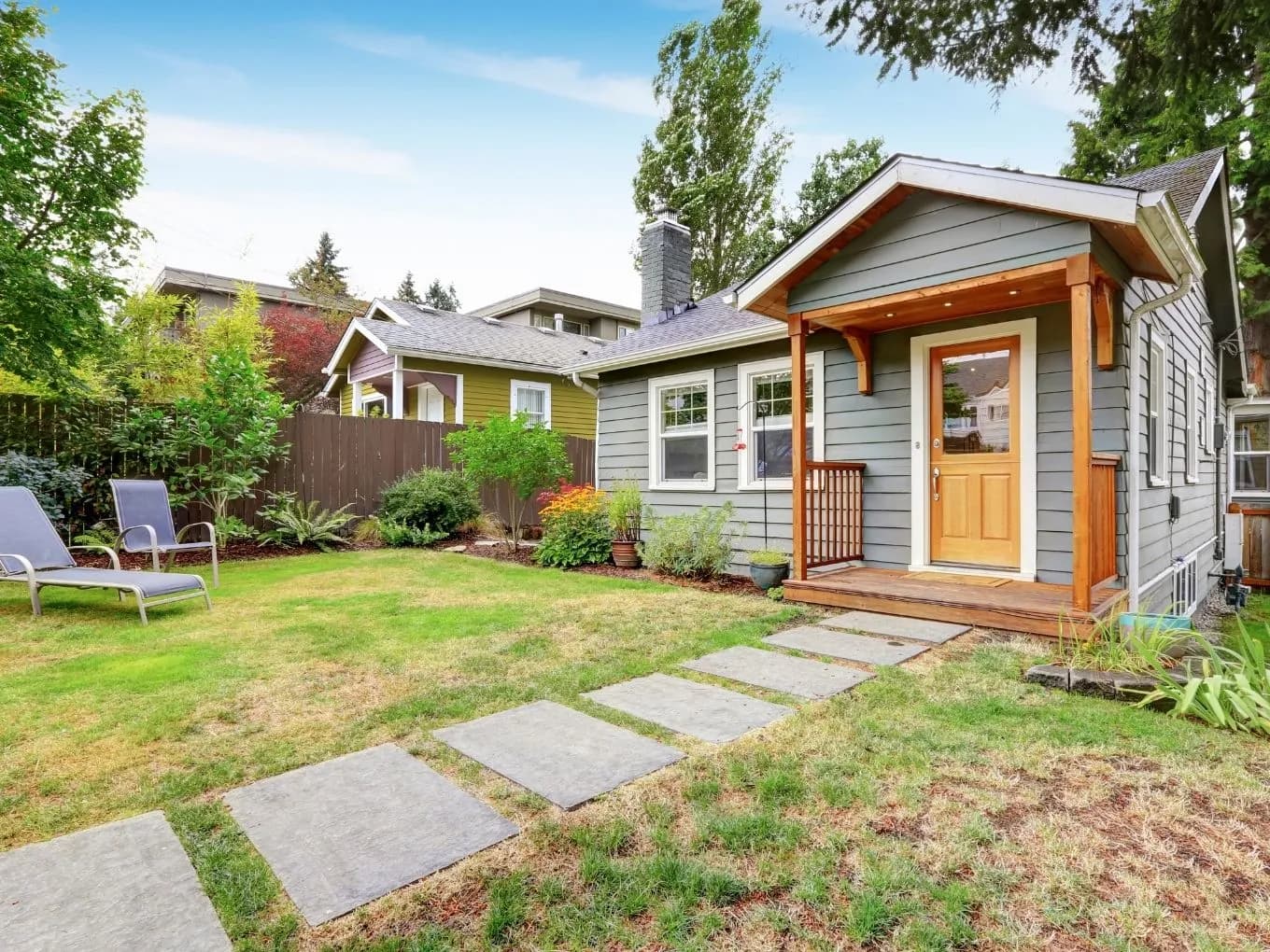 Backyard view of a small gray craftsman-style bungalow with wood-toned trim, a covered entry porch, and a natural wood front door with glass panels. Square concrete pavers form a path across the lawn to the porch steps, with two reclining patio chairs, garden plantings, and a wood privacy fence on the left. A larger olive-green house with a similar architectural style is visible next door under a mostly clear blue sky.