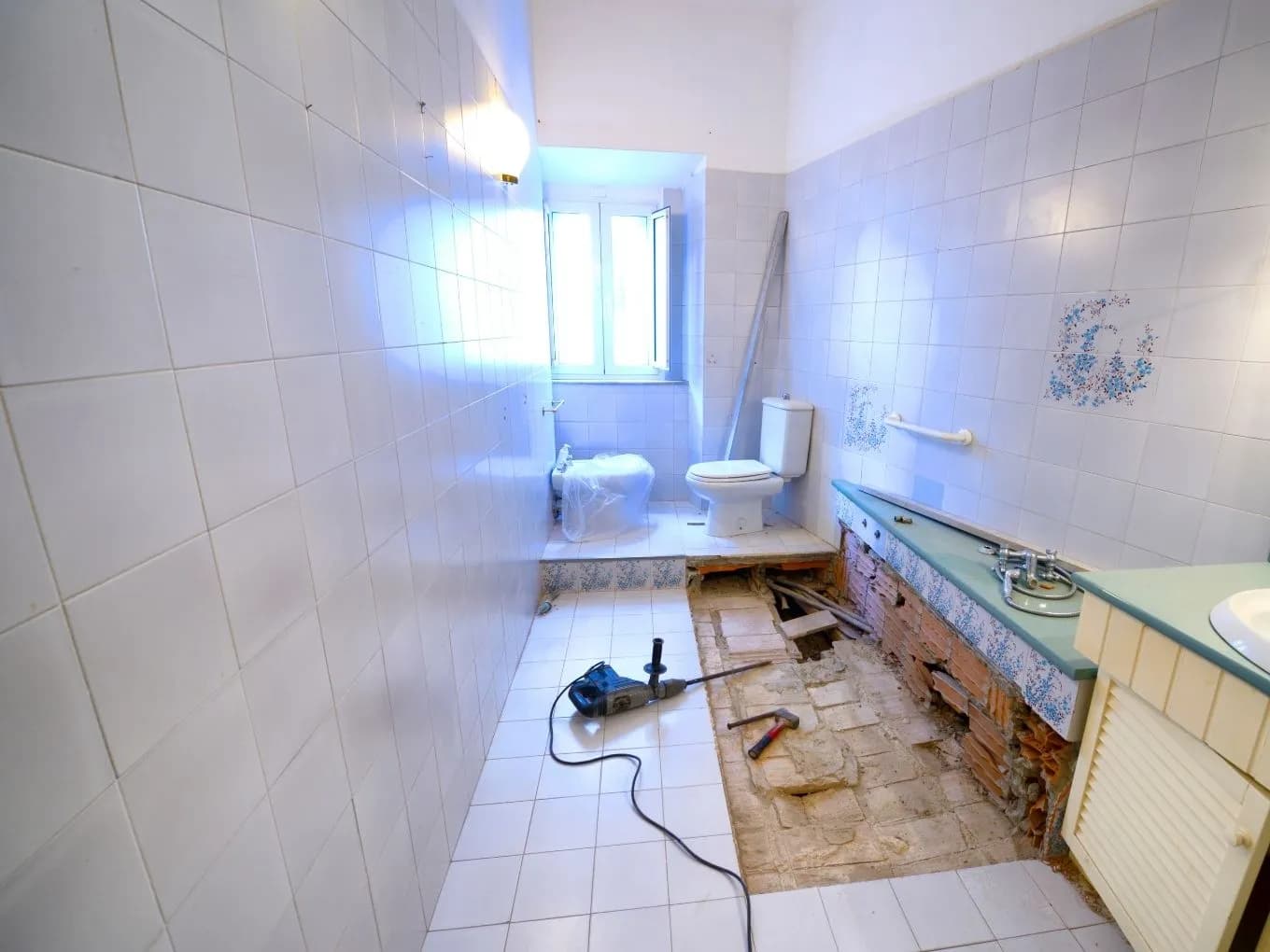 Interior of an outdated bathroom under renovation, fully tiled in white with decorative blue floral tile accents, showing an older toilet, a green countertop sink with exposed brick beneath it, and a section of floor tile removed to reveal brick and old plumbing pipes. A hammer drill with its cord and a hammer sit on the tiled floor, with sunlight streaming in through a small window and a wall-mounted grab bar visible near the sink.