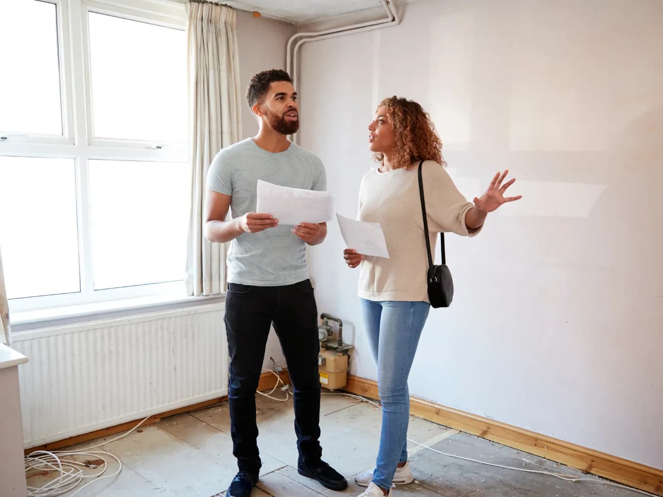 A man and woman stand talking in an empty, unfinished room with bare wood flooring and a wall that appears water-stained and unpainted, exposed pipes running along the ceiling. Both are holding papers, with the woman gesturing with one hand as if pointing out something about the room, suggesting they may be discussing a renovation or property viewing. A window with light curtains and an old radiator are visible on the left, with loose wiring on the floor.