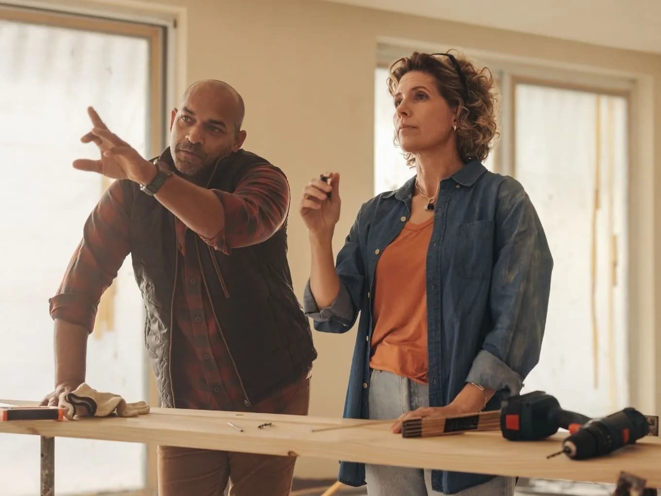 A man and woman stand at a wood workbench in a room with large windows, both gesturing upward as if discussing a plan or pointing out something in the room above them. A power drill, work gloves, pencils, and screws lie on the wooden plank in front of them, with soft natural light filtering through the windows in the background.
