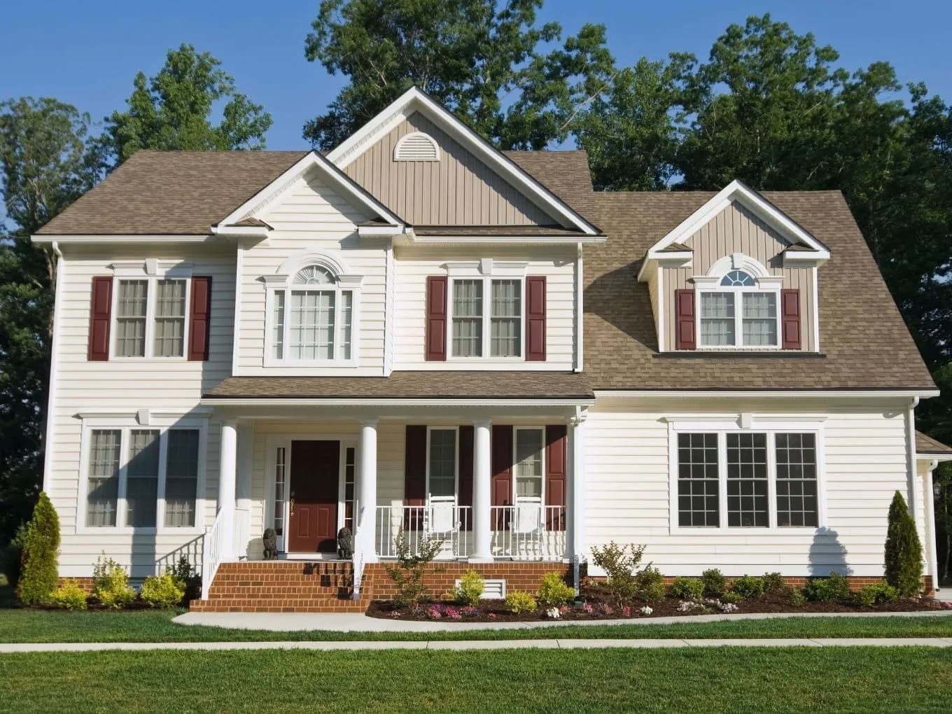 Exterior of a two-story cream-colored vinyl-sided house with dark red shutters and a brown shingled roof featuring multiple gables and a dormer window. A covered front porch with white columns and railings holds rocking chairs, and a red front door is accessed via brick steps flanked by small statues. Landscaped garden beds with flowering shrubs line the brick walkway and foundation, with a manicured lawn in the foreground and tall trees framing the house against a clear blue sky.