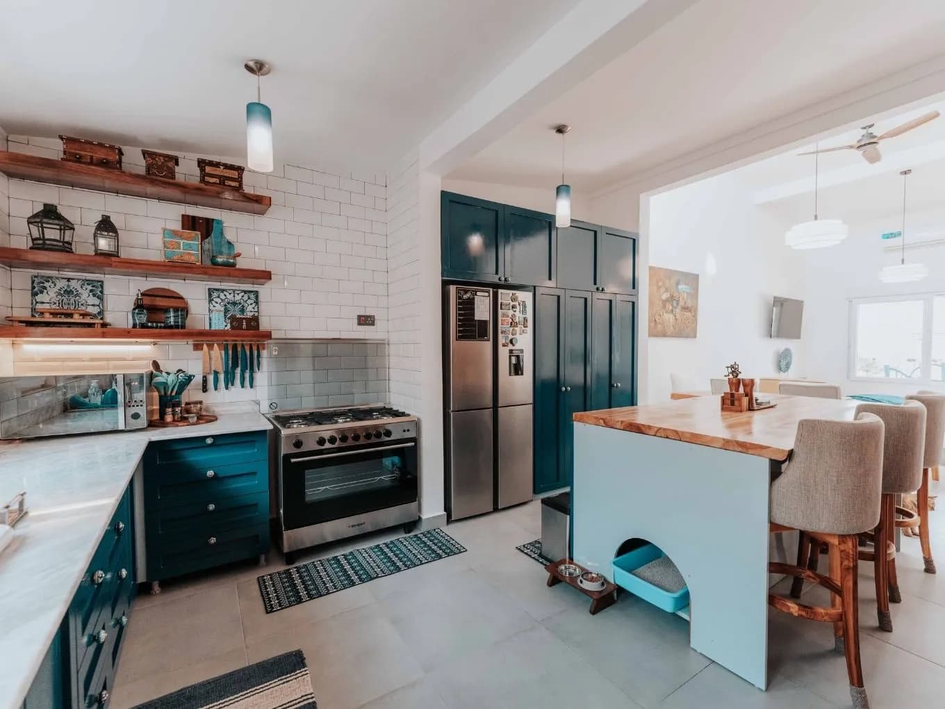 Colorful eclectic kitchen with teal cabinetry, white subway tile backsplash, and open wood shelves displaying decorative boxes, lanterns, and ceramic pieces. A stainless steel gas range and matching refrigerator sit against the wall, with a wood-topped kitchen island in a light blue base holding a built-in pet feeding area with a cat litter box underneath. The space opens into a bright adjoining dining area with upholstered chairs, a ceiling fan, and large windows in the background.