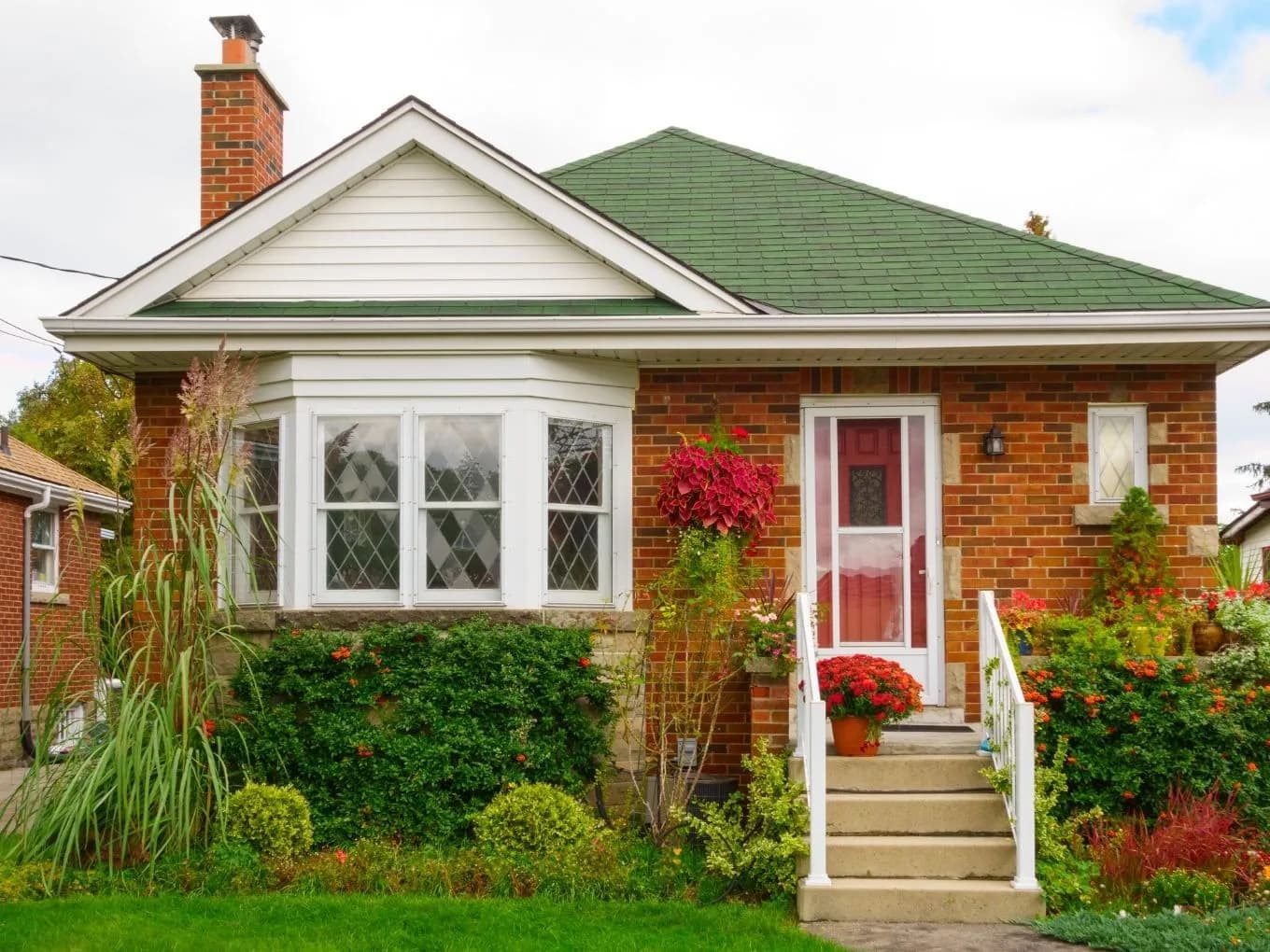 Exterior of a single-story red brick bungalow with a green shingled hip roof and a brick chimney. A white bay window with diamond-patterned panes sits below a white-trimmed front gable, and a red front door with a matching red storm door is flanked by white railings and concrete steps. Lush landscaping surrounds the entrance, including hanging red flowers, potted mums, ornamental grasses, and green shrubs, with a well-manicured lawn in the foreground.