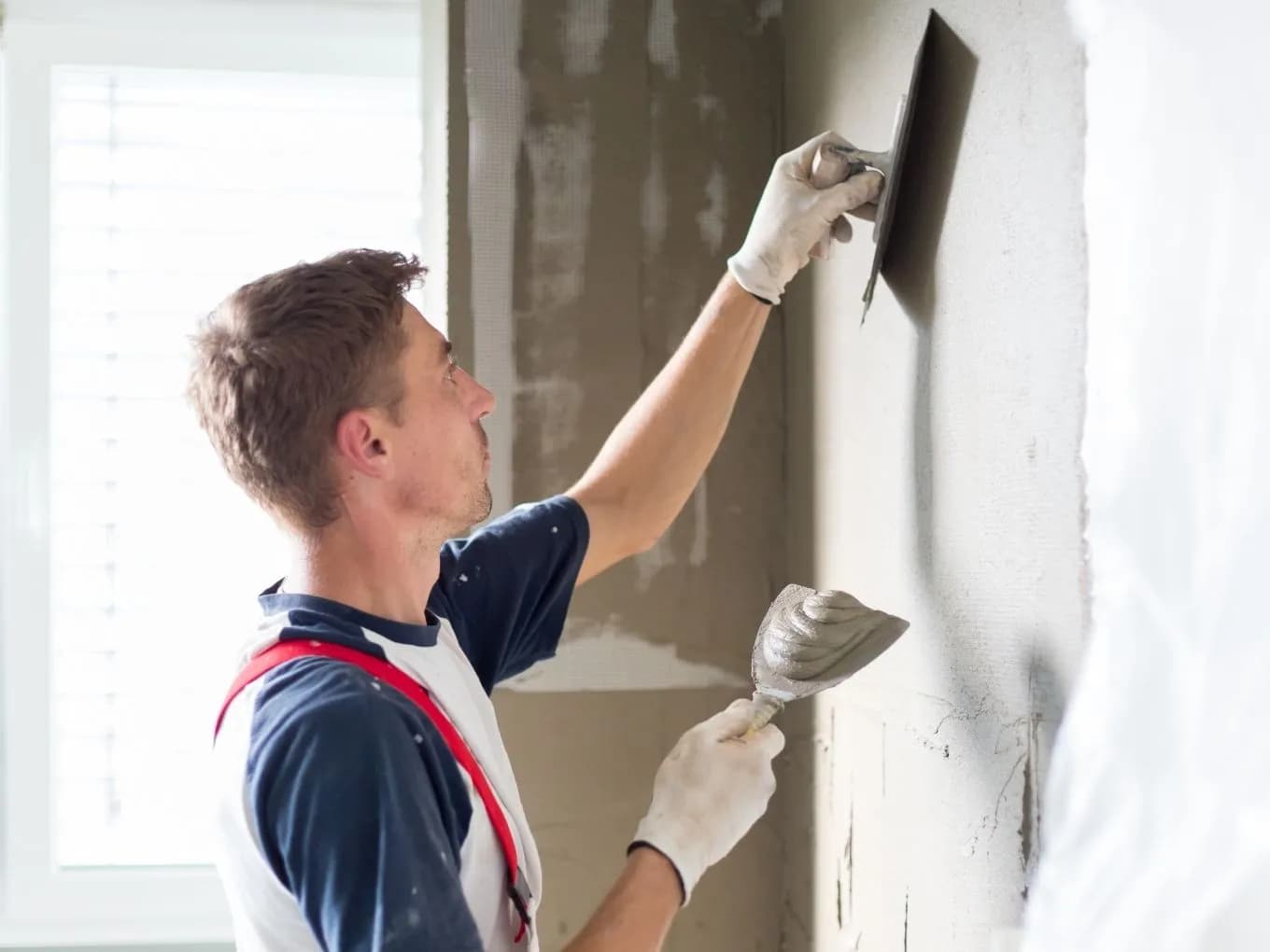 Close-up of a man wearing red suspenders and work gloves applying plaster to an interior wall, using a hawk to hold a mound of wet plaster in one hand and a trowel to smooth it onto the wall's mesh-reinforced surface with the other. A bright window with white shutters is visible in the background.