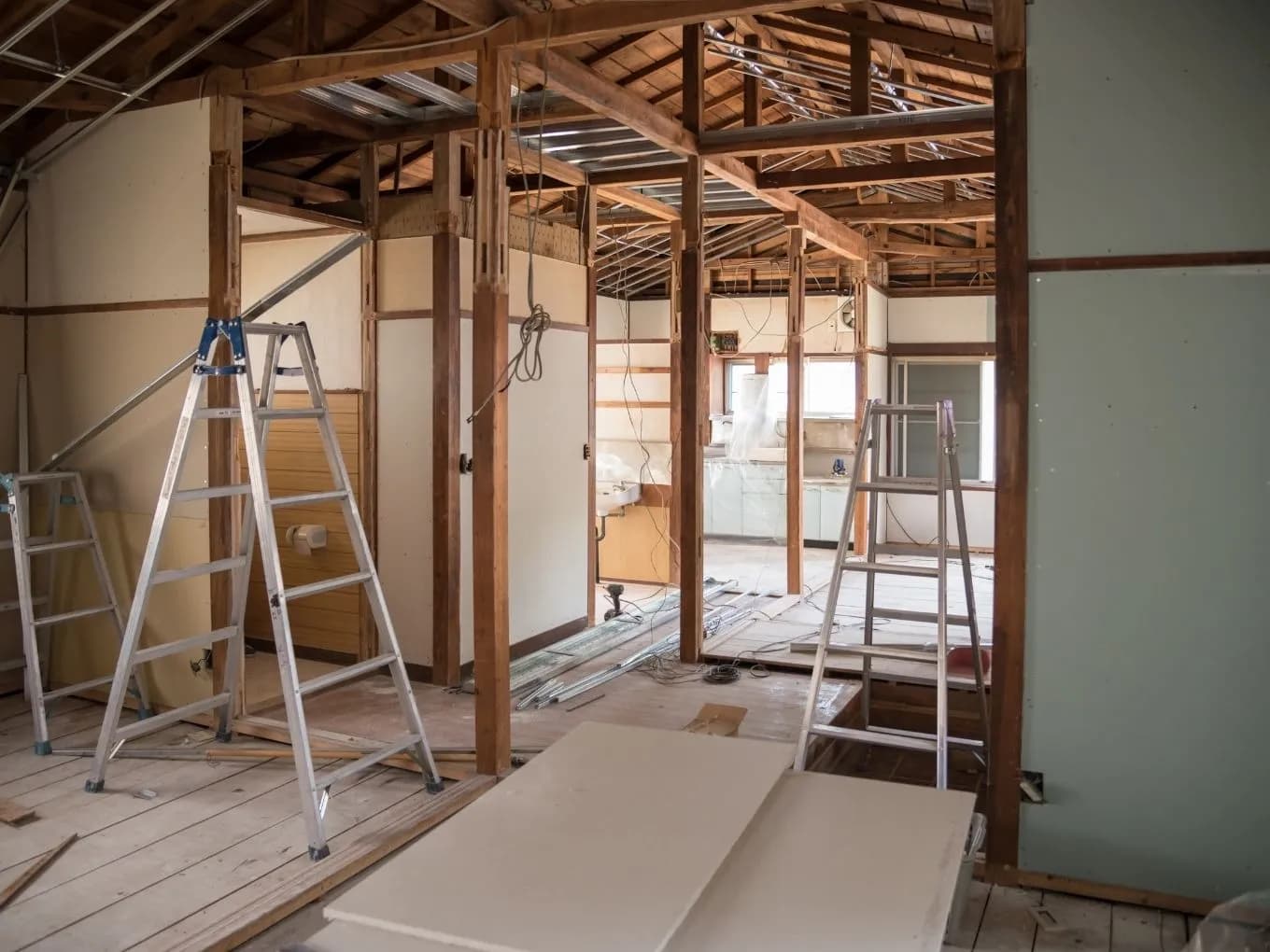 Interior of a traditional house mid-renovation, showing exposed wood beam framing, rafters, and structural posts with the ceiling and roof structure visible. Multiple aluminum step ladders stand on the wood plank floor amid loose wiring, drywall sheets, and construction debris. In the background, a partially demolished kitchen area is visible with cabinets covered in plastic sheeting and a window letting in natural light. traditional wood-framed doors remain on either side of the room.