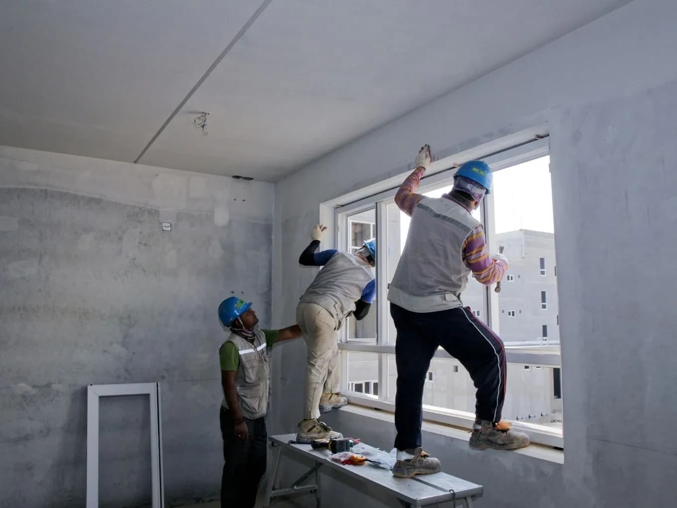 Three construction workers wearing blue safety helmets install a large window frame in an unfinished, gray-plastered room. Two workers stand on a metal folding table and along the window ledge to secure the top of the frame while a third supervises from the floor, with tools and a folded cloth resting on the table below. Additional multi-story buildings are visible through the window opening outside.