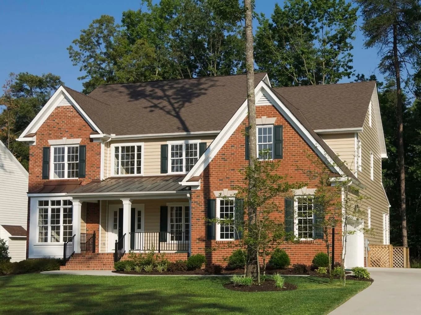 Exterior of a two-story red brick colonial-style house with tan siding accents, dark green shutters, and a metal standing-seam roof over the front porch. A white-columned entryway with black iron railings leads to the front door, and young trees and shrubs are planted in mulched beds around the manicured front lawn. Tall mature trees frame the house on either side under a clear blue sky, with a paved driveway visible on the right.