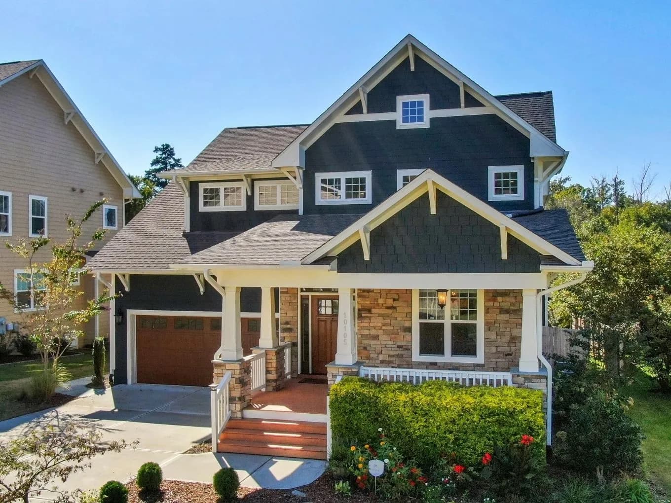 Exterior of a two-story Craftsman-style house with dark navy shingle siding, white trim, and decorative exposed rafter tails under multiple gables. A stone-clad front porch with white columns shelters a wood front door and leads down red-stained wood steps, with a two-car garage featuring wood-tone doors to the left. Landscaped shrubs, flowering plants, and a neighboring tan house are visible, with a concrete driveway in the foreground under a clear blue sky.